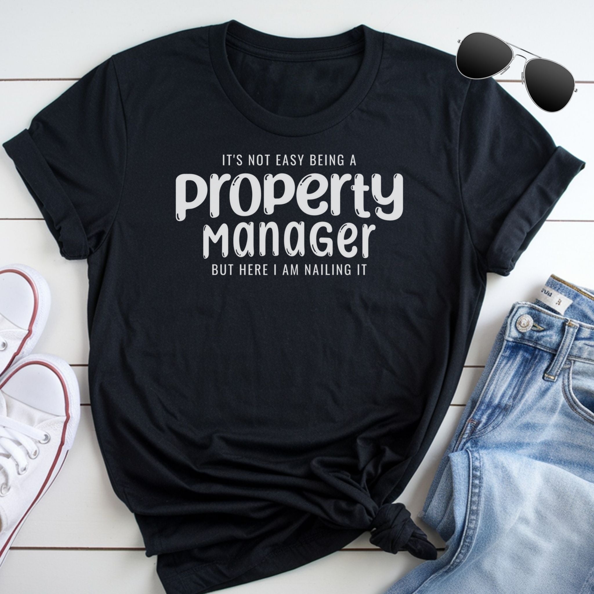 A black t-shirt with white text that reads "It's not easy being a Property Manager but here I am nailing it" is displayed on a white background, accompanied by a pair of sunglasses and a pair of jeans.