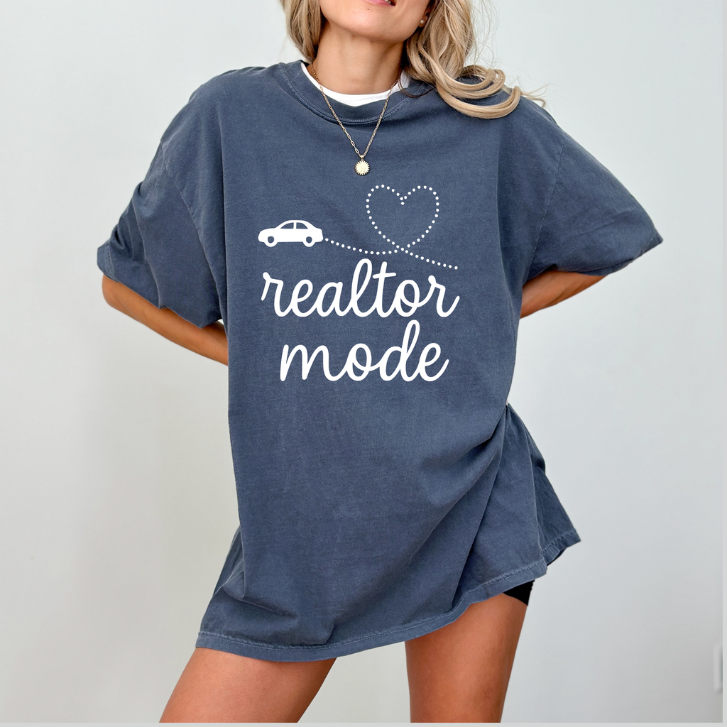 A woman wearing a blue t-shirt with the text "realtor mode" printed on it, standing against a white background.