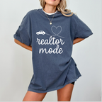 A woman wearing a blue t-shirt with the text "realtor mode" printed on it, standing against a white background.