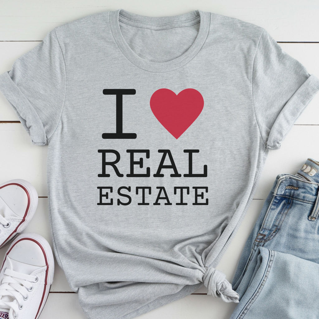 A gray t-shirt with the text "I ❤️ REAL ESTATE" printed on it, along with a red heart symbol. The t-shirt is laid out on a white surface, with a pair of white sneakers and a pair of blue jeans visible in the background.