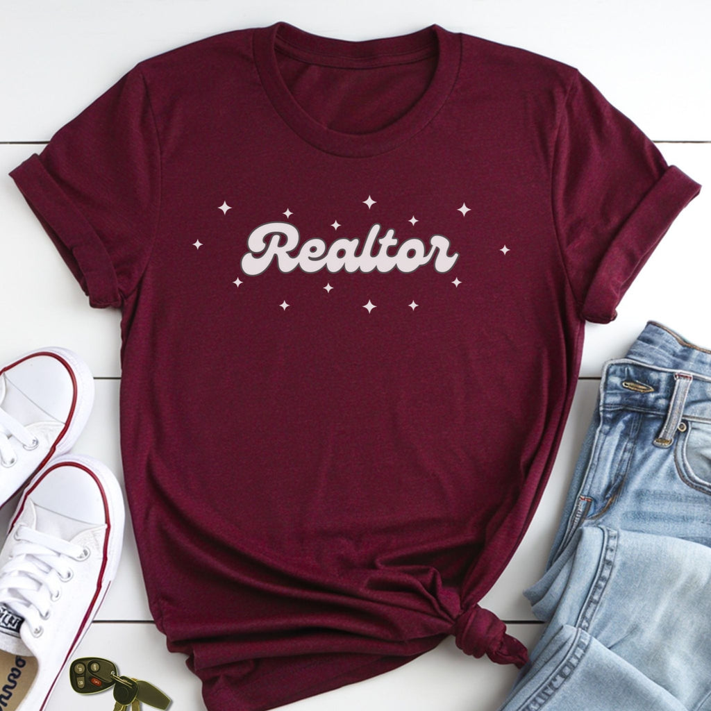A burgundy t-shirt with the word "Realtor" printed on it, along with a pair of jeans and white sneakers, all laid out on a white surface.