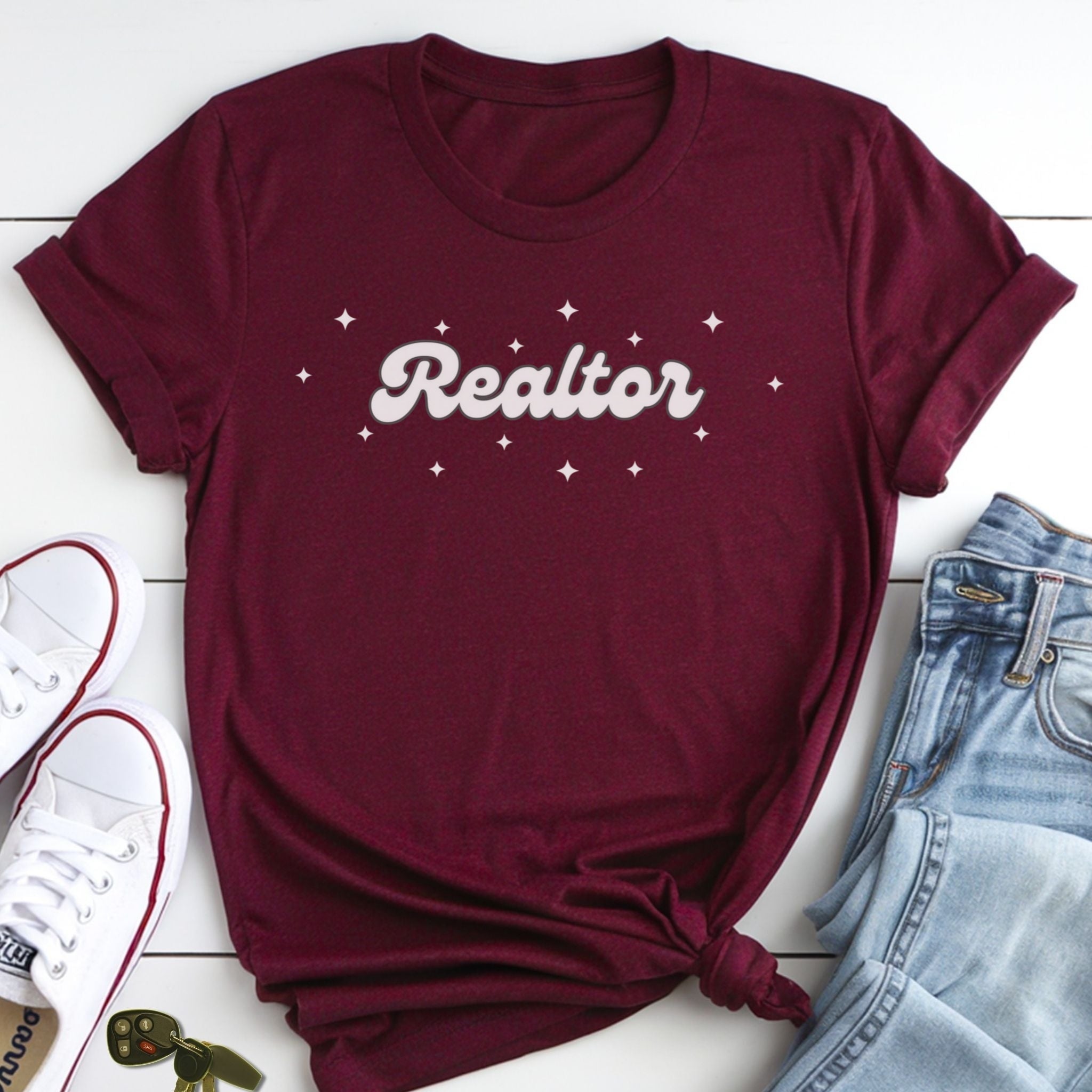 A burgundy t-shirt with the word "Realtor" printed on it, along with a pair of jeans and white sneakers, all laid out on a white surface.