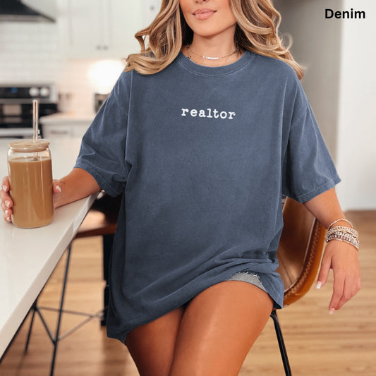 A woman wearing a blue t-shirt with the word "realator" printed on it, sitting at a kitchen counter with a drink in a glass.
