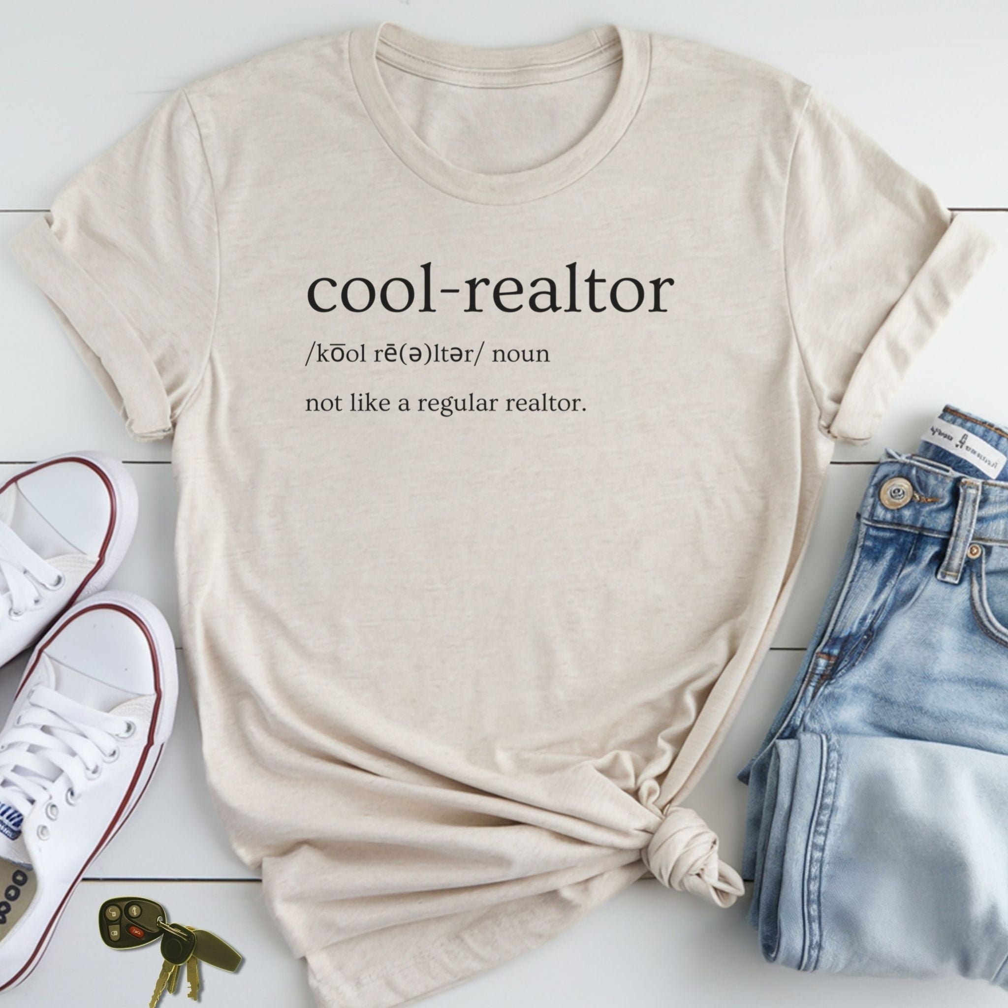 A beige t-shirt with the definition of a "cool-realtor" printed on it, along with a pair of jeans and sneakers, all laid out on a white surface.