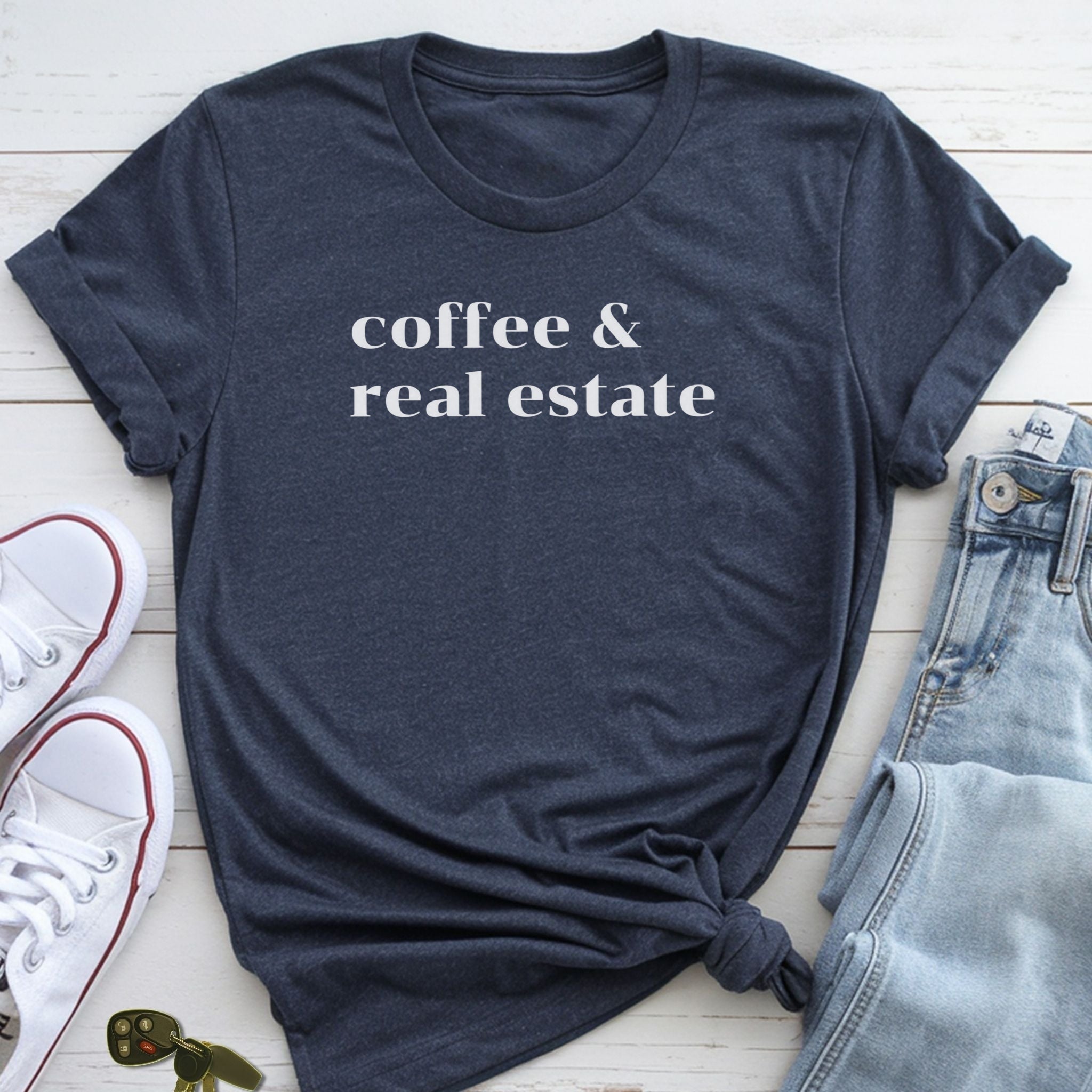 A navy blue t-shirt with the text "coffee & real estate" printed on it, along with a pair of jeans and white sneakers, all laid out on a white surface.
