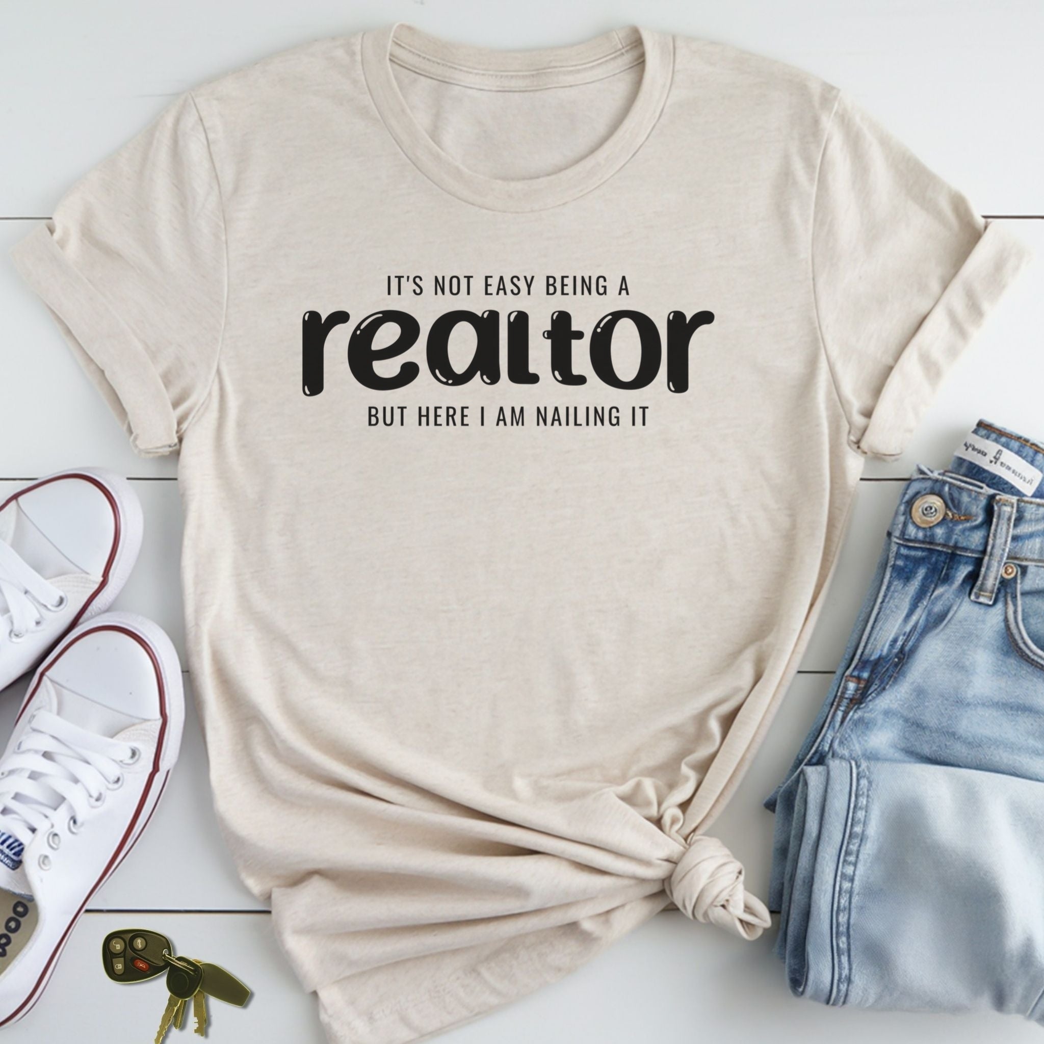 A beige t-shirt with the text "IT'S NOT EASY BEING A REALTOR BUT HERE I AM NAILING IT" printed on it, along with a pair of jeans and sneakers.