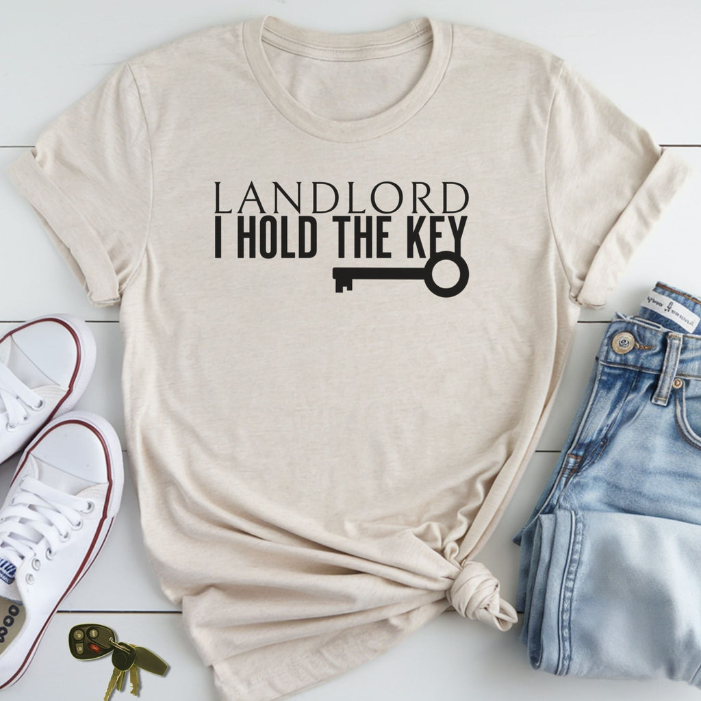 A beige t-shirt with the text "LANDLORD I HOLD THE KEY" printed on it, along with a key graphic. The t-shirt is placed on a white surface, accompanied by a pair of jeans and a pair of white sneakers.