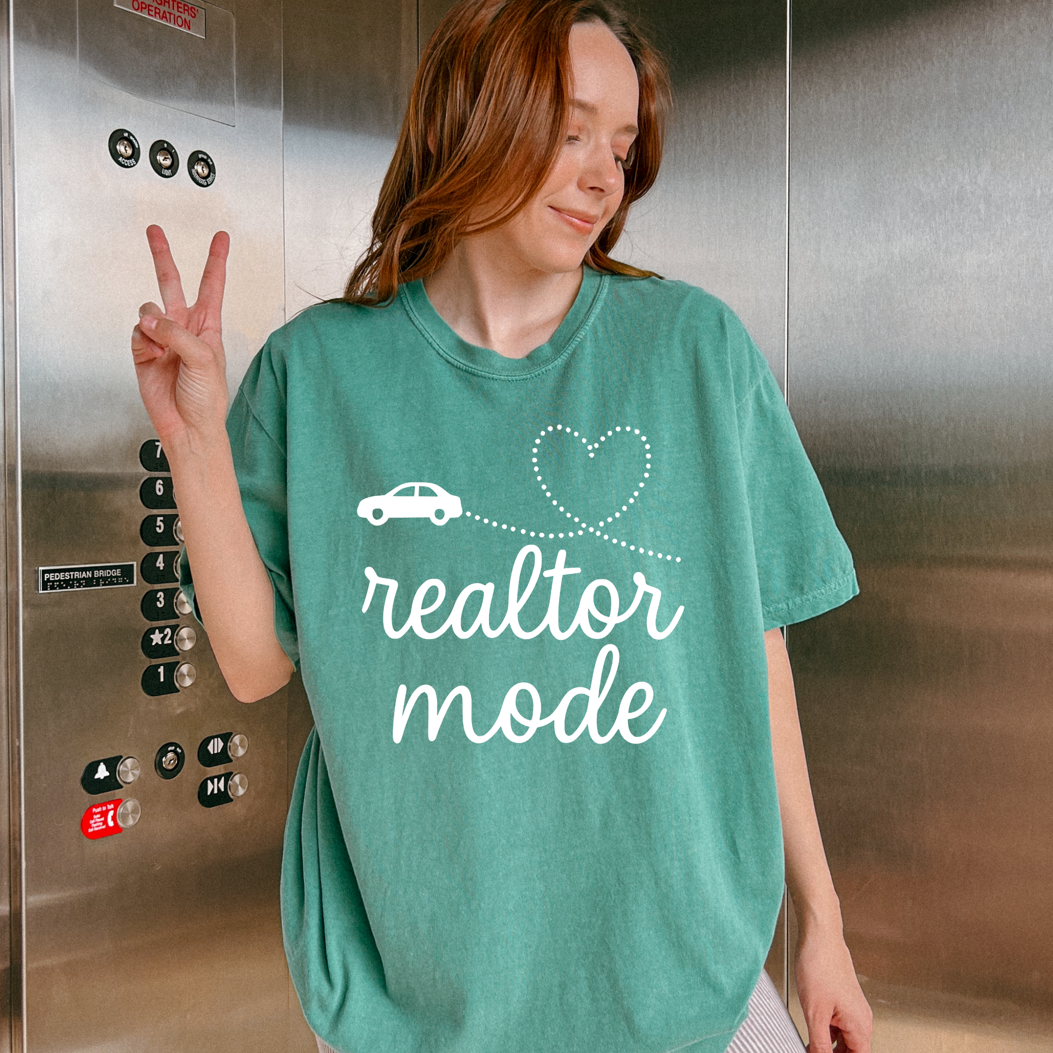 A woman wearing a green t-shirt with the text "realtor mode" and a heart symbol is standing in front of a metal elevator.