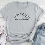 A gray t-shirt with the text "REAL ESTATE AGENT" printed on it, along with a simple graphic of a house. The t-shirt is laid out on a white surface, and there are a pair of jeans and white sneakers visible in the background.