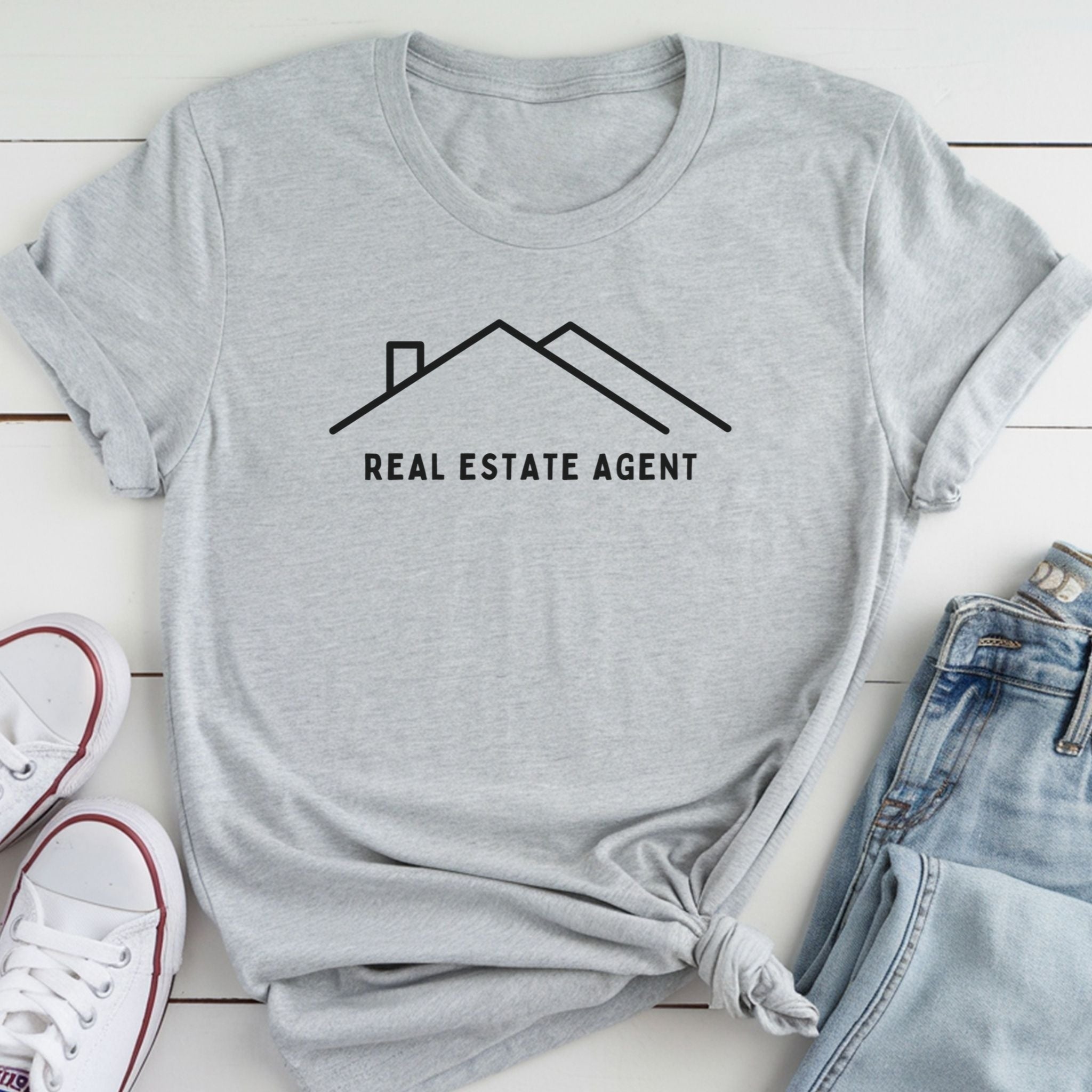 A gray t-shirt with the text "REAL ESTATE AGENT" printed on it, along with a simple graphic of a house. The t-shirt is laid out on a white surface, and there are a pair of jeans and white sneakers visible in the background.