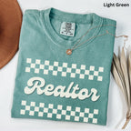 a light green t-shirt with the word "Realtor" printed on it, along with a checkered pattern. The t-shirt is placed on a white surface, and there is a brown hat and some dried leaves nearby.