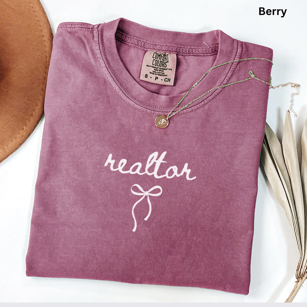 A pink t-shirt with the word "realtor" printed on it, along with a necklace and a brown hat, all placed on a white surface.