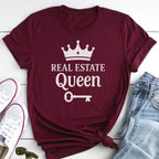 A burgundy t-shirt with the text "REAL ESTATE QUEEN" printed on it, along with a crown and a key, is displayed on a white surface alongside a pair of jeans and white sneakers.