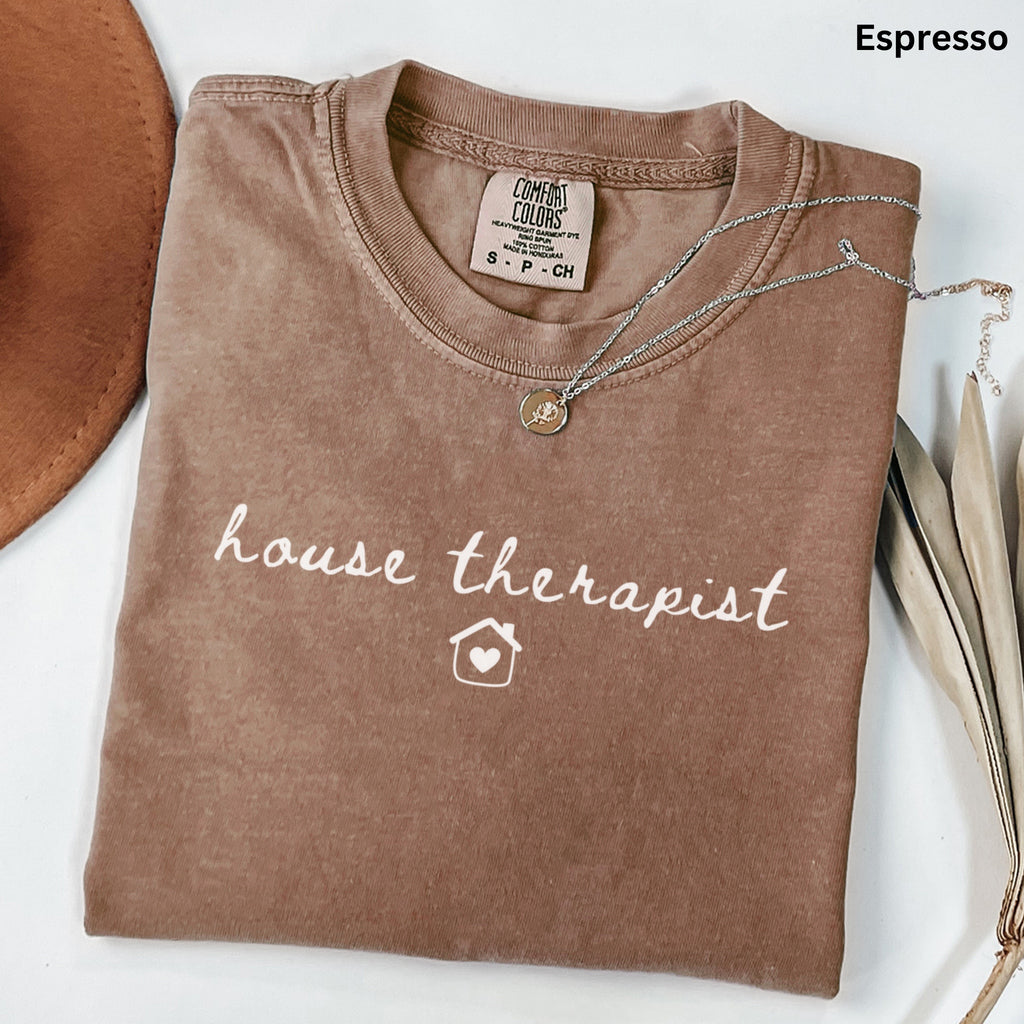 a brown t-shirt with the text "house therapist" printed on it, along with a small house icon. The t-shirt is laid out on a white surface, and there is a necklace and some leaves nearby.