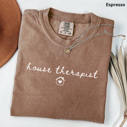 a brown t-shirt with the text "house therapist" printed on it, along with a small house icon. The t-shirt is laid out on a white surface, and there is a necklace and some leaves nearby.