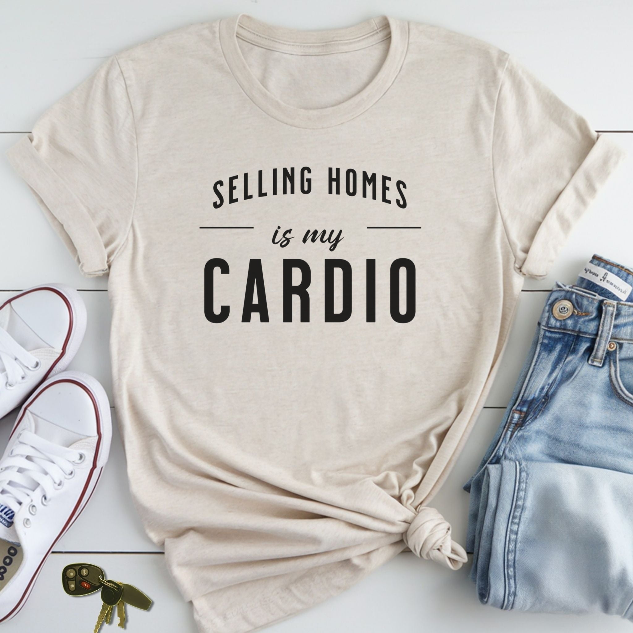 A beige t-shirt with the text "Selling homes is my cardio" printed on it, along with a pair of jeans and sneakers.