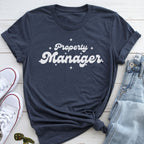 A navy blue t-shirt with the text "Property Manager" printed on it, along with some jeans and white sneakers, all laid out on a wooden surface.
