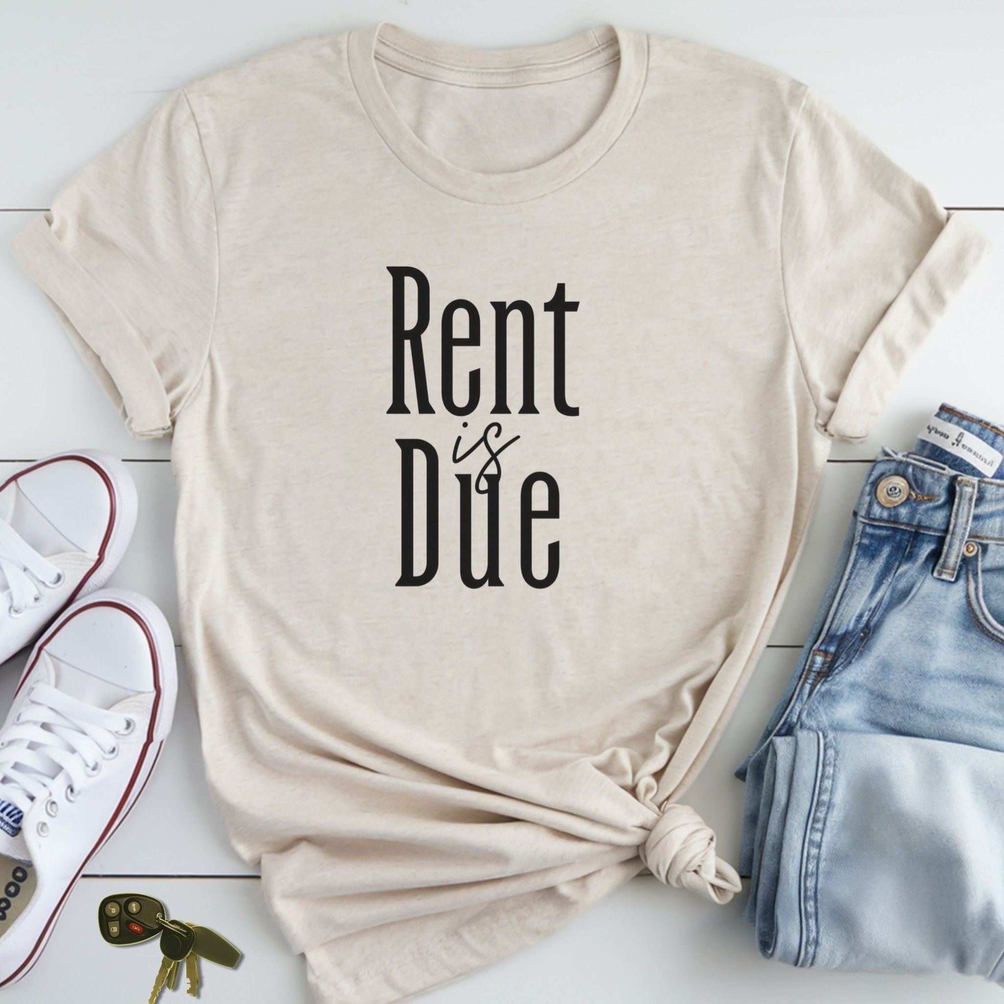 A beige t-shirt with the text "Rent is Due" printed on it, along with a pair of jeans and sneakers, all laid out on a white surface.