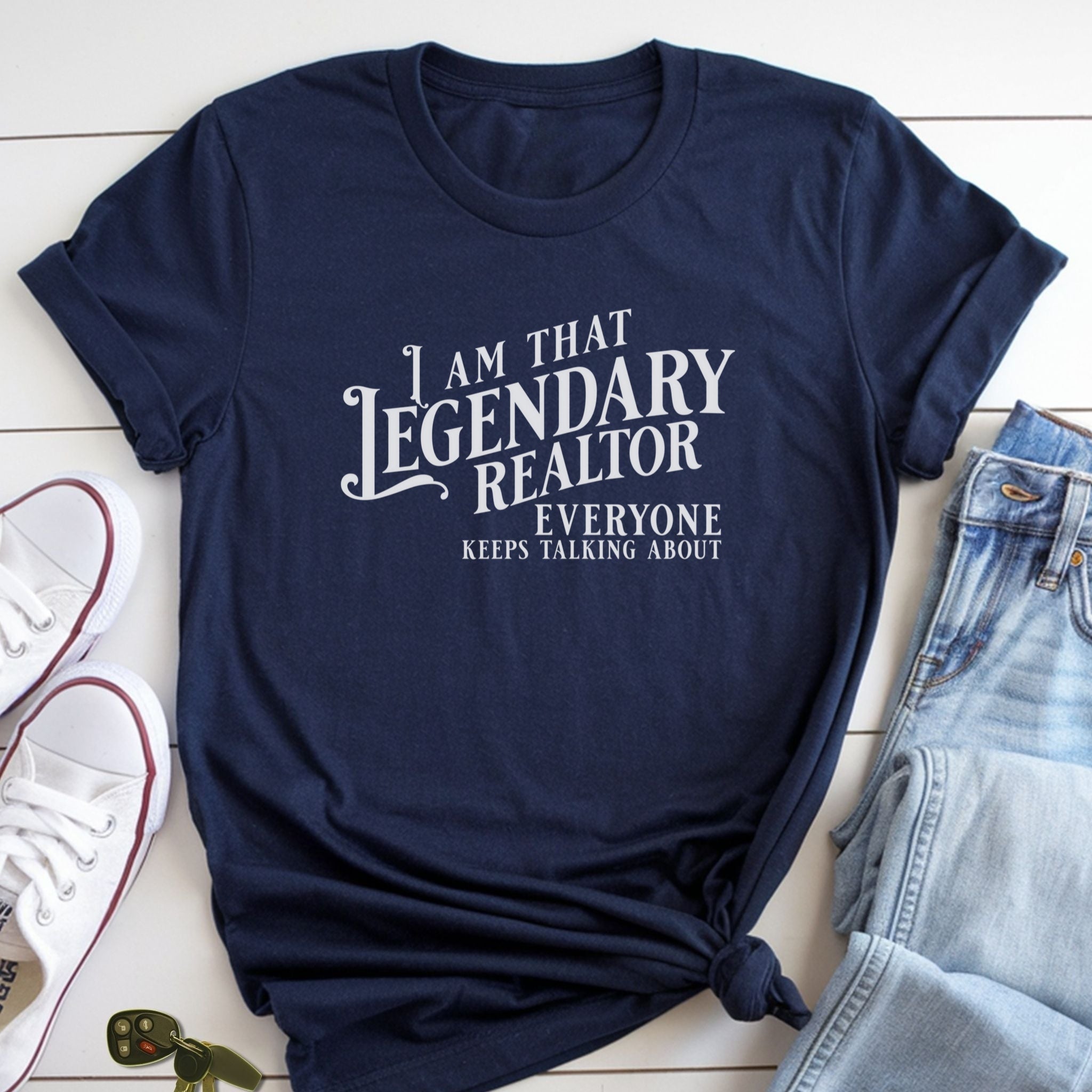 A navy blue t-shirt with white text that says "I am that Legendary Realtor everyone keeps talking about" is laid out on a white surface, accompanied by a pair of jeans and a pair of white sneakers.