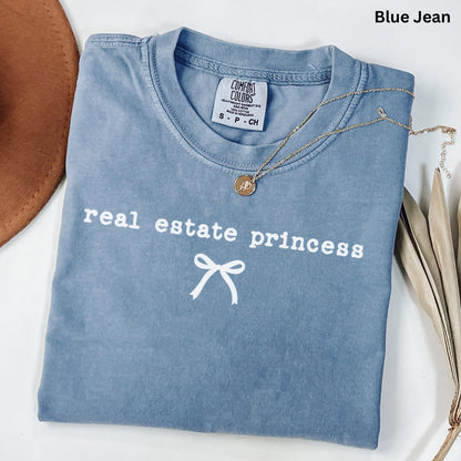 A blue t-shirt with the text "real estate princess" printed on it, along with a ribbon design. The t-shirt is laid out on a white surface, and there is a necklace and some leaves nearby.
