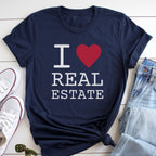 A navy blue t-shirt with the text "I ♥ REAL ESTATE" printed on it, along with a red heart symbol. The t-shirt is laid out on a white surface, with a pair of jeans and white sneakers visible in the background.