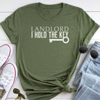 A green t-shirt with the text "LANDLORD I HOLD THE KEY" printed on it, along with a key graphic. The t-shirt is placed on a white surface, and there are a pair of sunglasses, a pair of sneakers, and a pair of jeans nearby.
