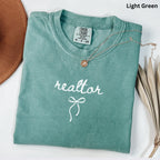 A light green t-shirt with the word "realtor" printed on it, along with a small gold pendant necklace. The t-shirt is folded and placed on a white surface, with a brown hat and some dried leaves visible in the background.