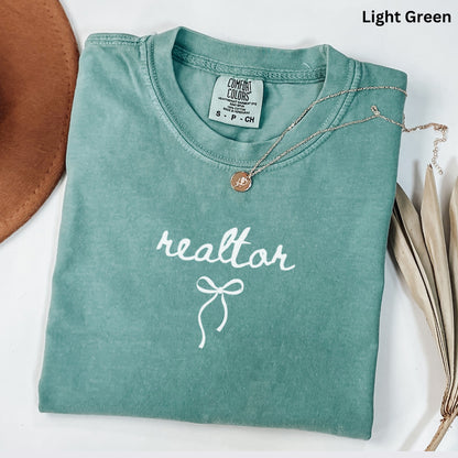 A light green t-shirt with the word "realtor" printed on it, along with a small gold pendant necklace. The t-shirt is folded and placed on a white surface, with a brown hat and some dried leaves visible in the background.