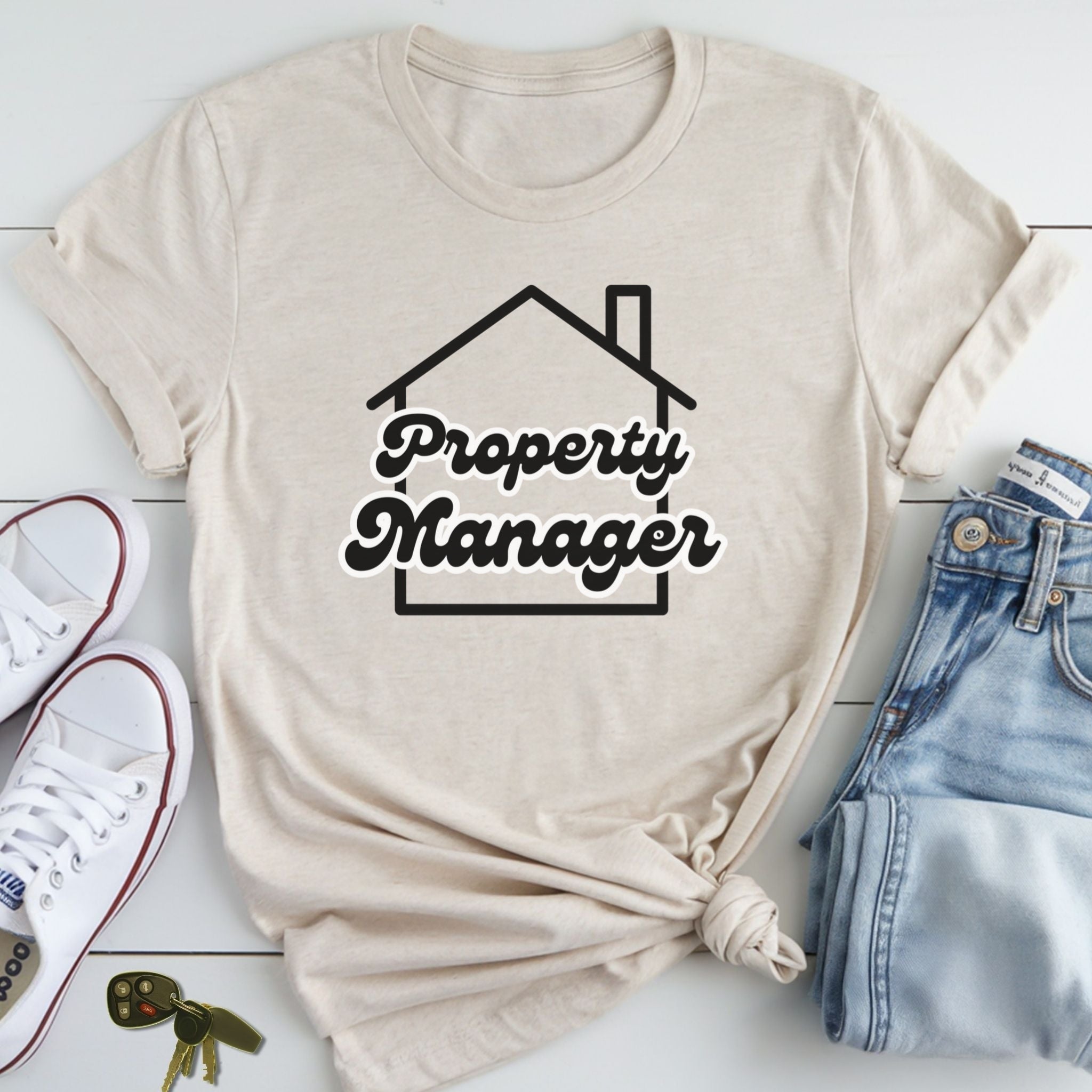 A beige t-shirt with the text "Property Manager" printed on it, along with a graphic of a house, is laid out on a white surface next to a pair of jeans and sneakers.
