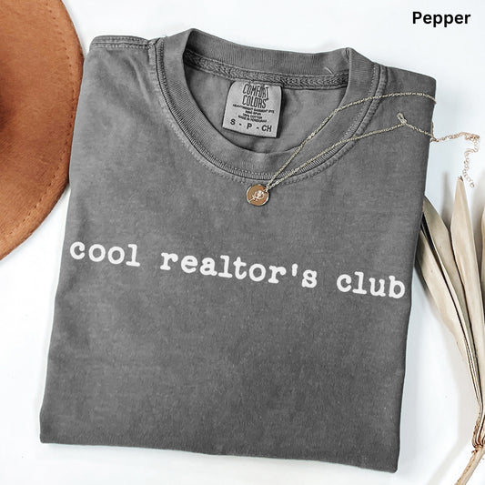 A gray t-shirt with the text "cool realtor's club" printed on it, along with a small coin-like pendant. The t-shirt is folded and placed on a white surface, with a brown hat and some leaves visible in the background.