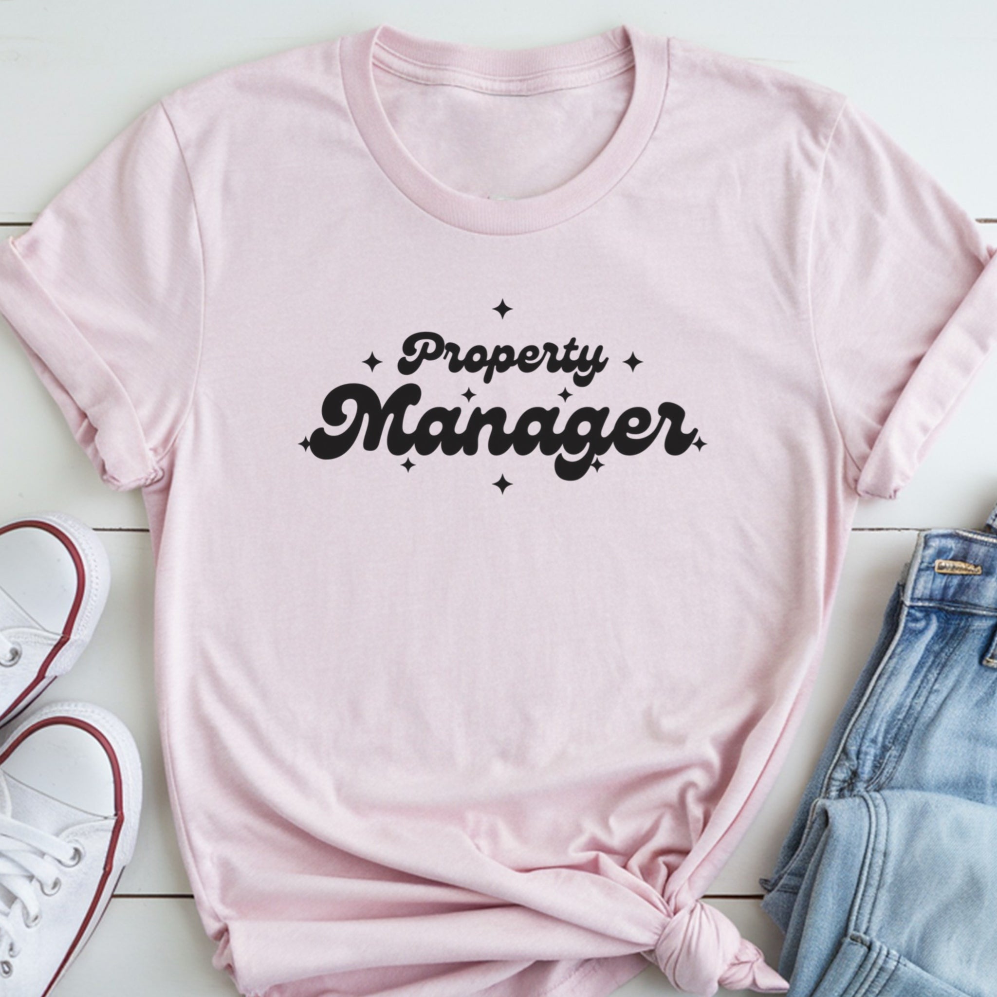 A pink t-shirt with the text "Property Manager" printed on it, along with some stars, is displayed on a white surface next to a pair of jeans and white sneakers.